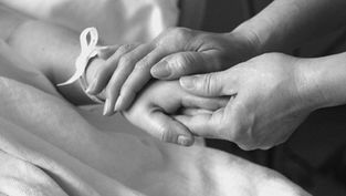 Hands gently holding another's in a comforting gesture. A ribbon is tied around one wrist. Monochrome image conveys warmth and care.