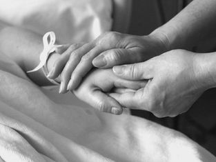 Hands gently holding another's in a comforting gesture. A ribbon is tied around one wrist. Monochrome image conveys warmth and care.