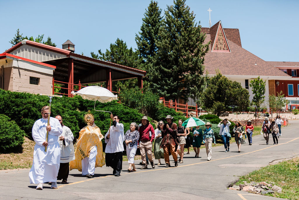 Religious procession of clergy and parishioners walking along a road by a church, carrying umbrellas and banners on a sunny day
