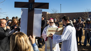 People in religious attire gather outside around a large cross on a sunny day. One person reads from a book, while others observe solemnly.