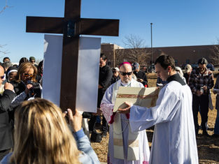 People in religious attire gather outside around a large cross on a sunny day. One person reads from a book, while others observe solemnly.