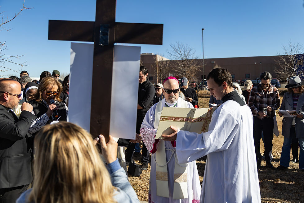 People in religious attire gather outside around a large cross on a sunny day. One person reads from a book, while others observe solemnly.