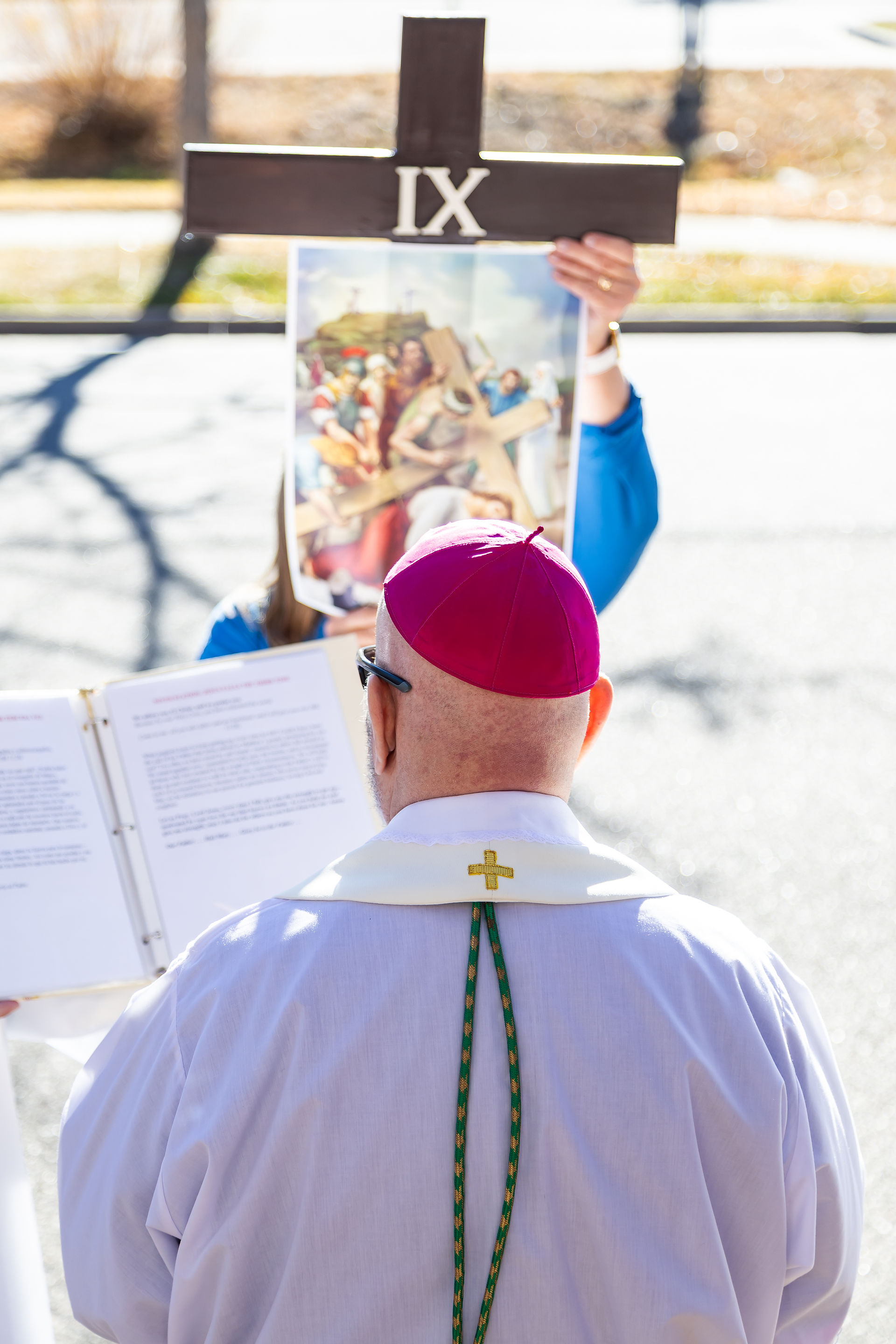 A clergyman in a white robe reads a book outdoors. A person holds a cross with "IX" and a painting of a biblical scene in the background.