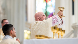 Bishop in ornate robes holds up a gold chalice during a church ceremony, with stained glass in the background. Solemn atmosphere.