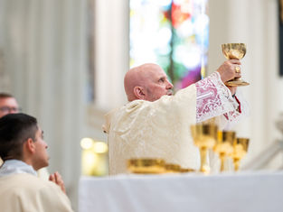 Bishop in ornate robes holds up a gold chalice during a church ceremony, with stained glass in the background. Solemn atmosphere.