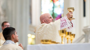 Bishop in ornate robes holds up a gold chalice during a church ceremony, with stained glass in the background. Solemn atmosphere.