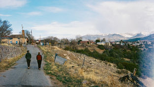 Two people walk on a rural road with mountains in the background. The setting is calm, with scattered houses and dry grass.