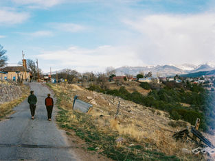 Two people walk on a rural road with mountains in the background. The setting is calm, with scattered houses and dry grass.