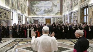 A religious leader addresses a group in an ornate hall with frescoes. Attendees in robes and suits, some clapping, in a solemn atmosphere.