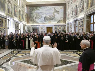 A religious leader addresses a group in an ornate hall with frescoes. Attendees in robes and suits, some clapping, in a solemn atmosphere.