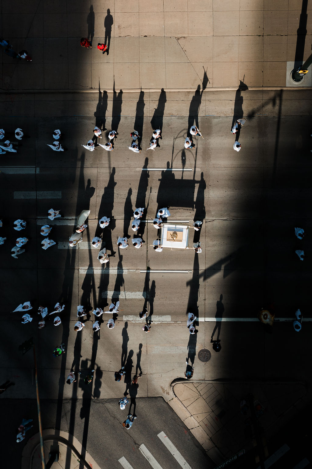 Aerial view of marchers in white on a city street, casting long shadows across the pavement in bright sunlight.