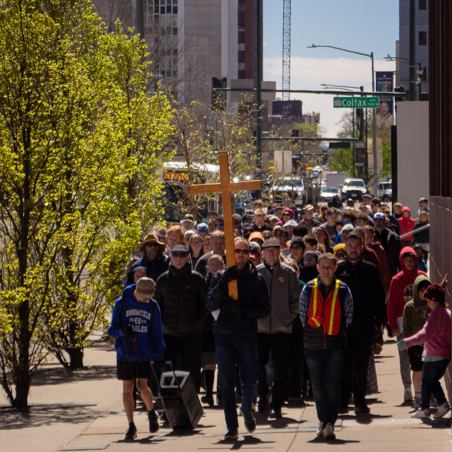 A large group of participants walk along a city sidewalk behind a wooden cross during a public religious procession.