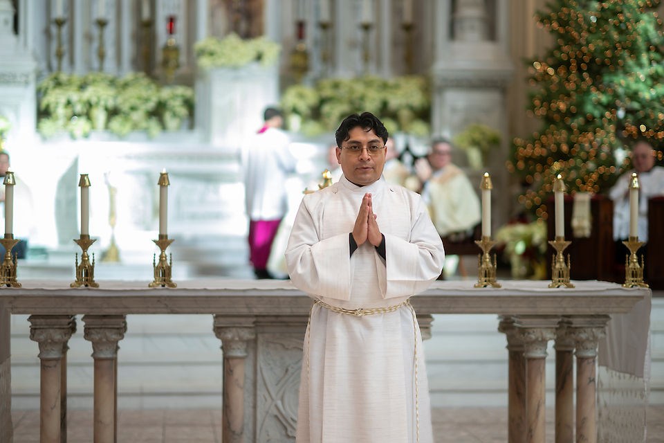 Man in white robe prays at altar with golden candlesticks. Background: ornate church, Christmas tree with lights, people blurred. Calm mood.