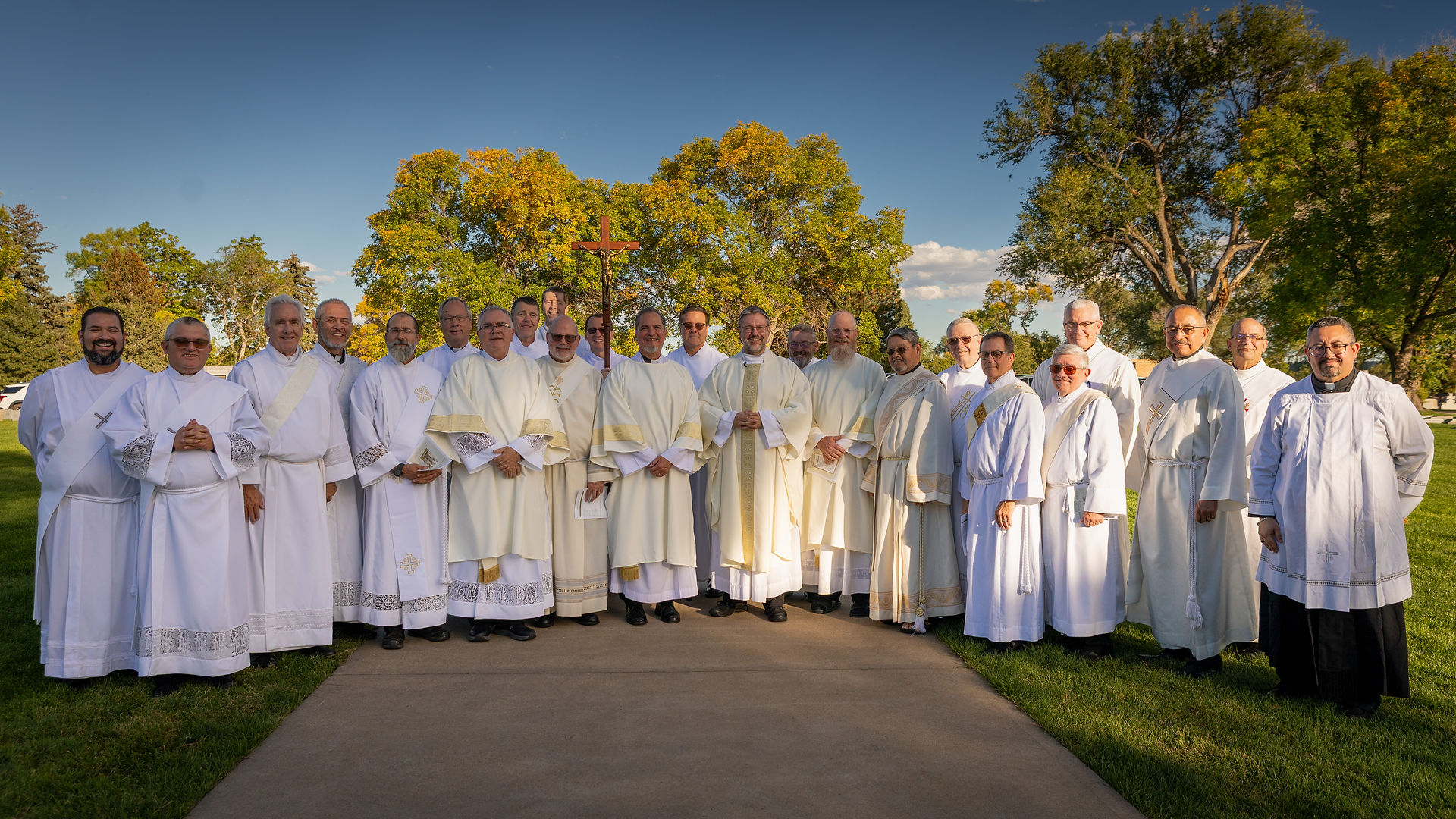 A group of clergy in white robes stands outdoors on a sunny day, smiling. Green trees and a blue sky are in the background.