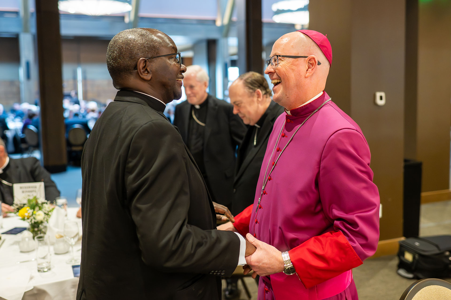 A bishop in magenta clerical attire smiles and shakes hands with a bishop in a black suit during a reception, with other clergy and guests in the background.