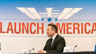 Man in a suit sits at a table with a "Launch America" backdrop. Two nameplates, one labeled Elon Musk and a NASA logo, are visible.