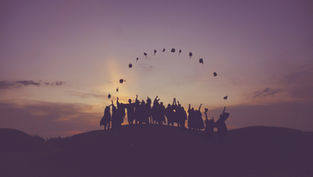 Silhouetted graduates joyfully toss caps into the purple-orange sunset sky, standing on a hill, celebrating together in a desert scene.