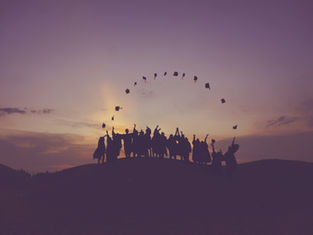 Silhouetted graduates joyfully toss caps into the purple-orange sunset sky, standing on a hill, celebrating together in a desert scene.