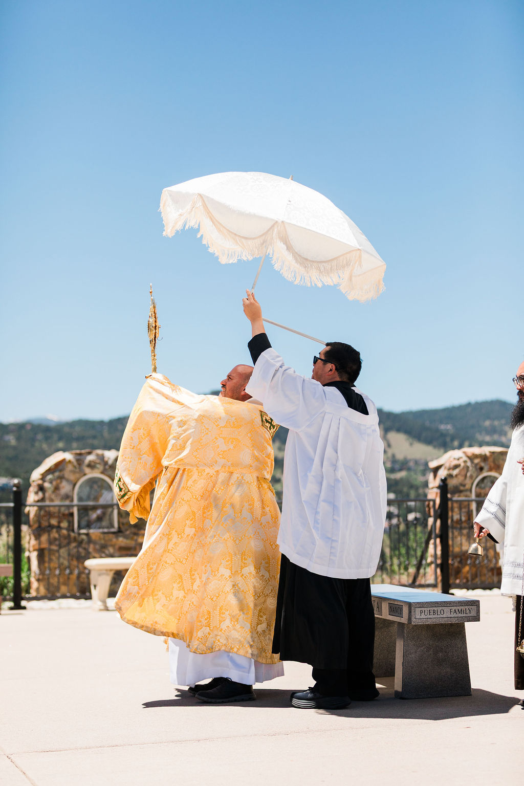 Two priests in robes perform an outdoor ritual, one holding a white fringed umbrella; a bell and Pueblo Family bench appear by mountains.