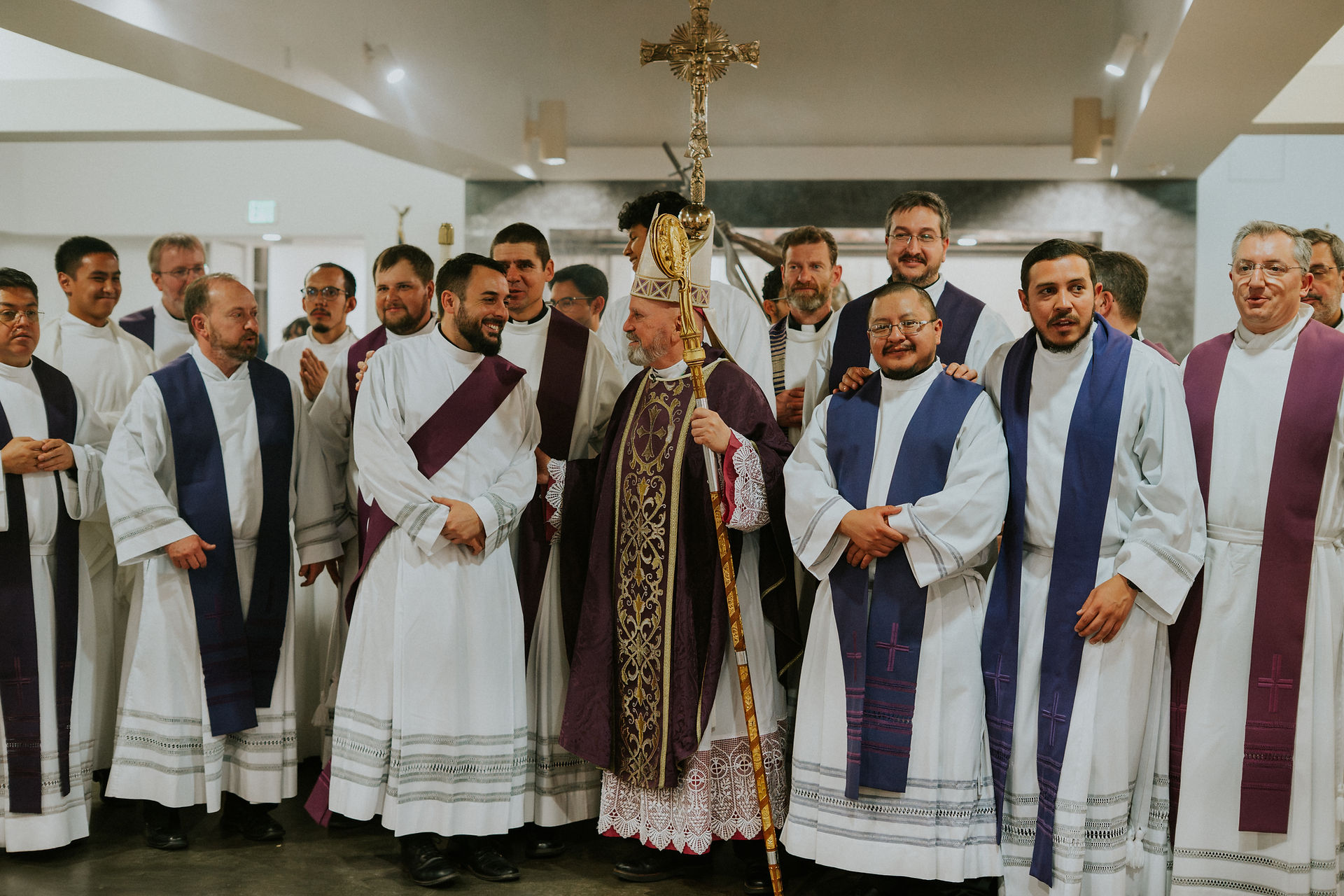 A group of clergy, including a bishop holding a crosier, pose together indoors. They wear white and purple robes, smiling warmly.