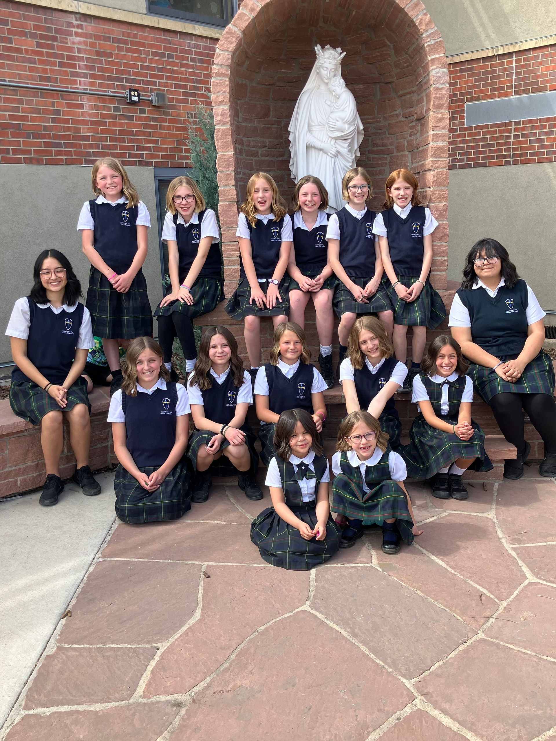 Group of schoolgirls in uniform pose on steps in front of a statue of the Virgin Mary holding the Child Jesus.