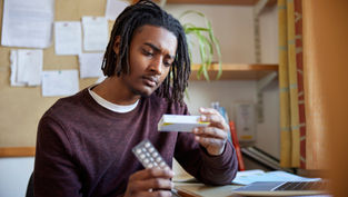 Man in a burgundy sweater examines medicine packaging and blister pack at a desk with a laptop, books, and plants in the background. Thoughtful mood.