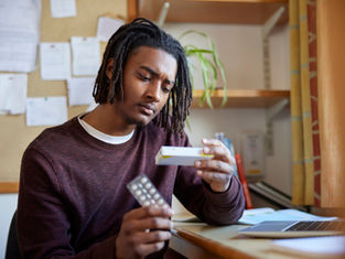 Man in a burgundy sweater examines medicine packaging and blister pack at a desk with a laptop, books, and plants in the background. Thoughtful mood.