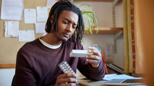 Man in a burgundy sweater examines medicine packaging and blister pack at a desk with a laptop, books, and plants in the background. Thoughtful mood.