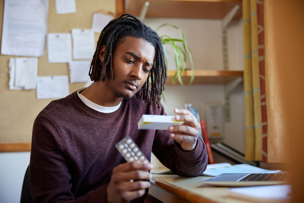 Man in a burgundy sweater examines medicine packaging and blister pack at a desk with a laptop, books, and plants in the background. Thoughtful mood.