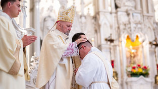 A bishop in ceremonial attire blesses a kneeling man in white robes inside an ornately decorated church, with a standing man praying nearby.