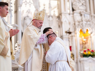 A bishop in ceremonial attire blesses a kneeling man in white robes inside an ornately decorated church, with a standing man praying nearby.