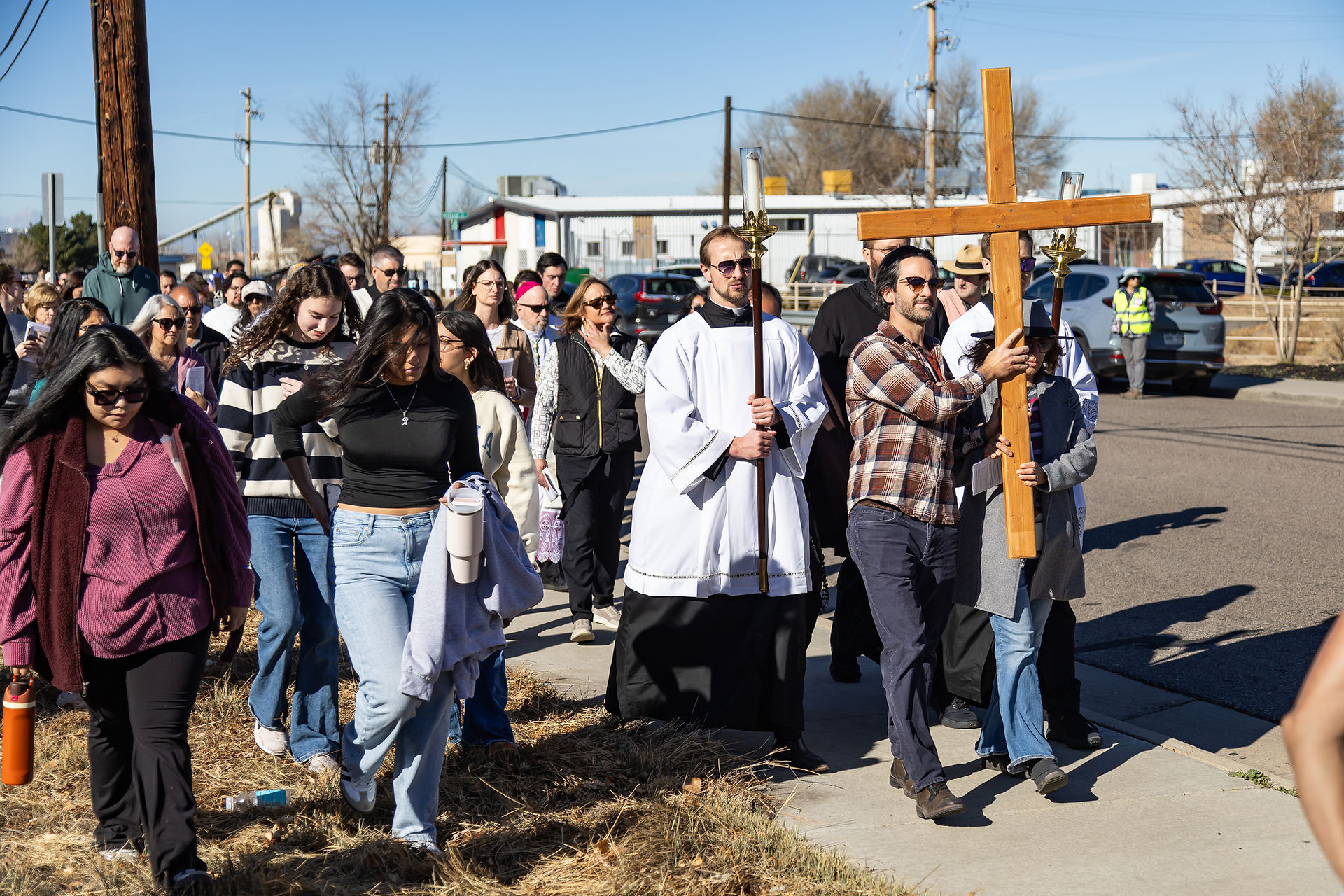 A group of people, some in religious attire, walk along a street holding a large wooden cross under a clear blue sky.