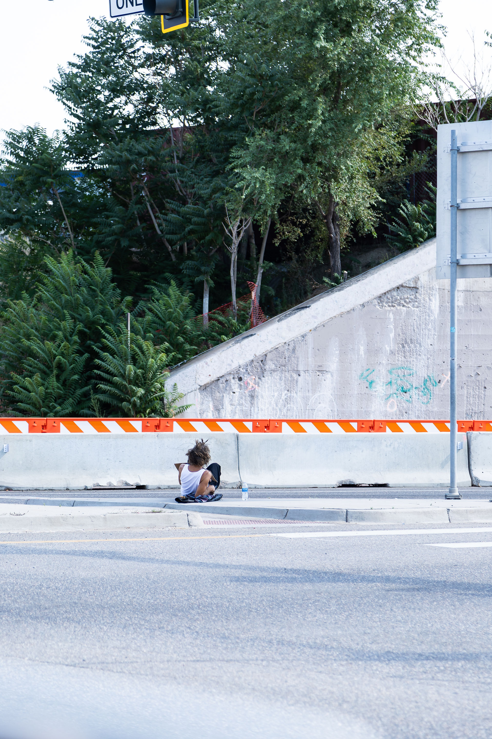 Person sitting on the sidewalk by a concrete barrier with orange stripes, near a road and trees. A bottle of water is beside them.