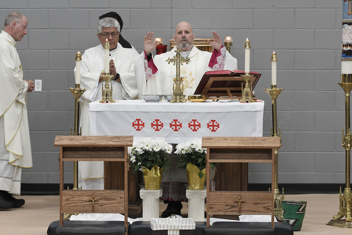 Priest conducting a mass at an altar with candles, cross, and open book. Assistants stand nearby. Gray brick wall background.