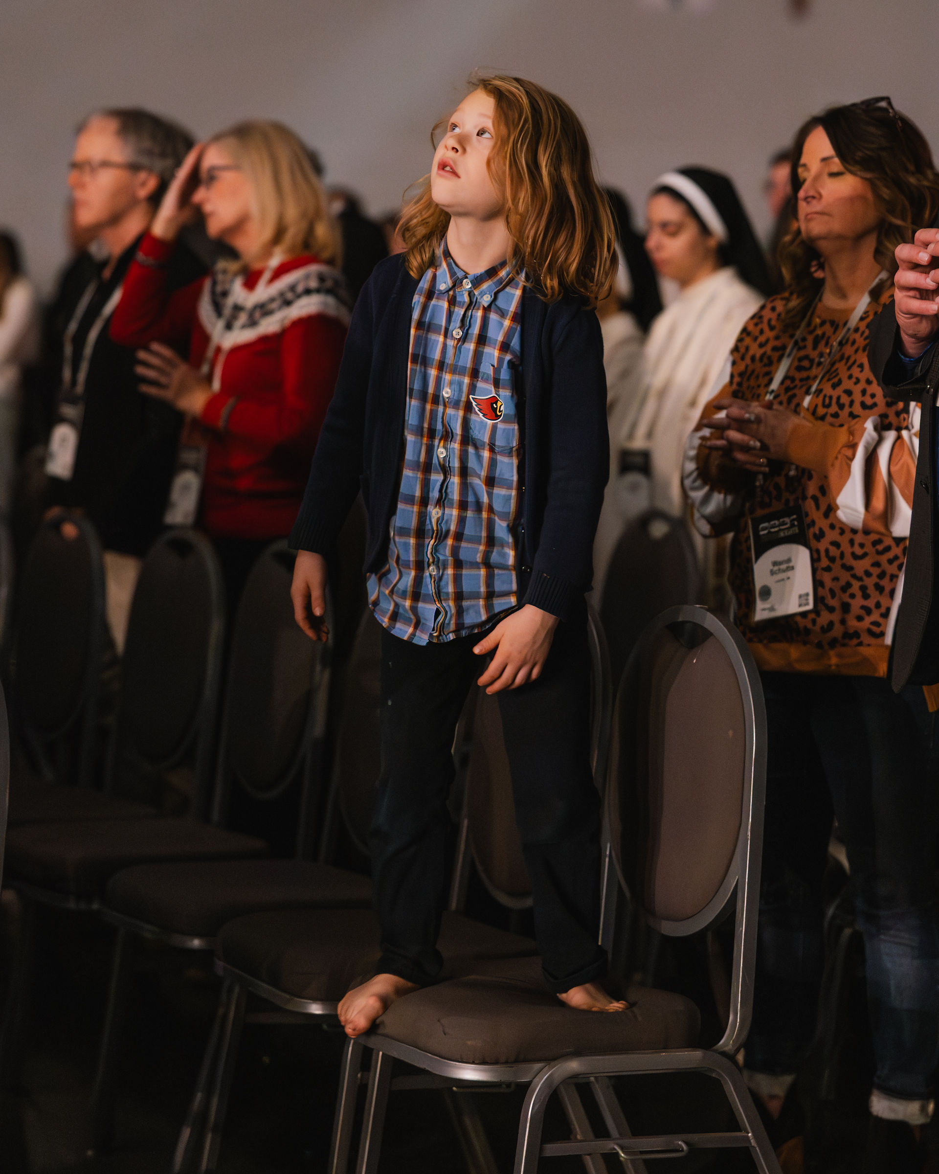 Young boy in a plaid shirt stands barefoot on a chair in a crowded room, looking upward. People around him appear focused and attentive.