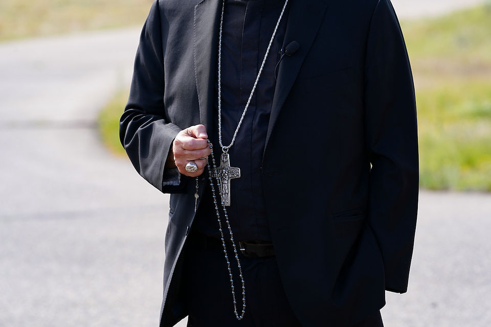 Clergyman in black suit holds a rosary with cross outdoors on a road, calm and solemn.