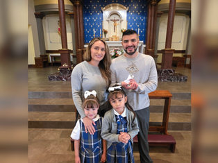 A family of four stands in a church. Parents in gray, daughters in plaid uniforms. The father holds a religious item. Blue altar backdrop.