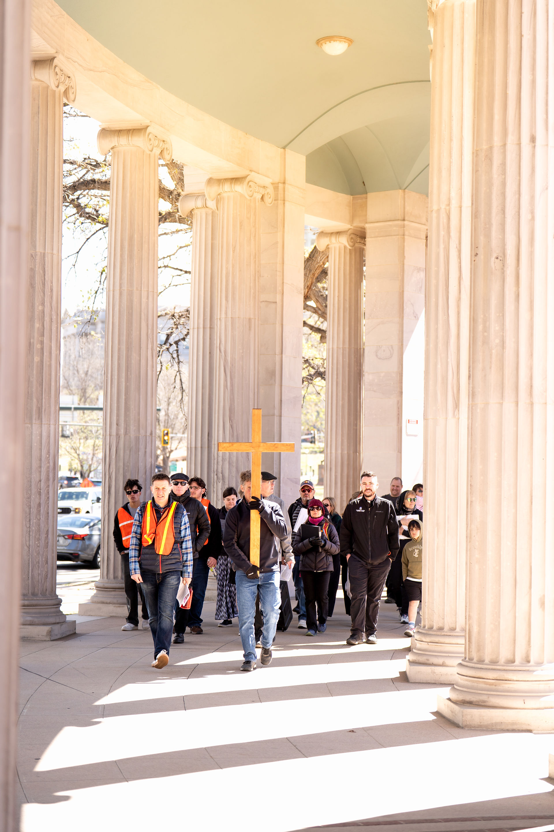 A group walks beneath a colonnade, following a wooden cross during a public Way of the Cross procession.