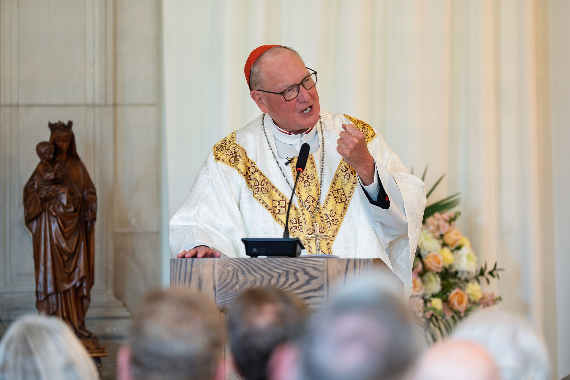 A religious leader in ornate robes speaks passionately at a wooden podium. A statue and floral arrangement are in the background.