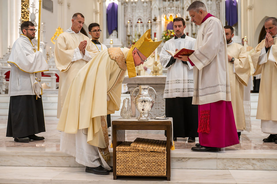 A bishop in ceremonial attire bows before a table with ceremonial objects in a church, surrounded by clergy in white robes, solemn atmosphere.