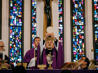 Bishop holds up a host during Mass, surrounded by clergy in purple vestments. Stained glass windows in the background create a solemn setting.
