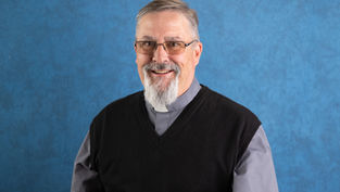 Smiling man with glasses and a gray clerical shirt, wearing a black sweater. Blue textured background. Appears calm and approachable.