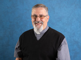Smiling man with glasses and a gray clerical shirt, wearing a black sweater. Blue textured background. Appears calm and approachable.