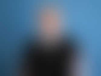 Smiling man with glasses and a gray clerical shirt, wearing a black sweater. Blue textured background. Appears calm and approachable.