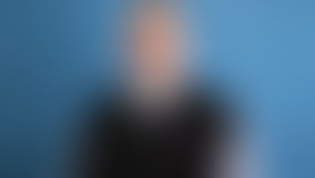 Smiling man with glasses and a gray clerical shirt, wearing a black sweater. Blue textured background. Appears calm and approachable.