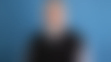 Smiling man with glasses and a gray clerical shirt, wearing a black sweater. Blue textured background. Appears calm and approachable.
