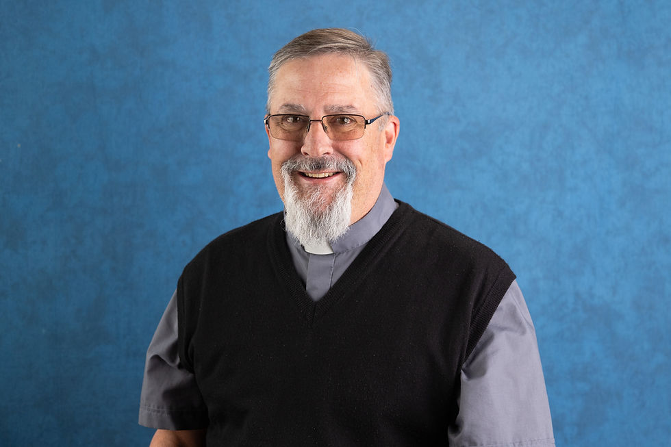 Smiling man with glasses and a gray clerical shirt, wearing a black sweater. Blue textured background. Appears calm and approachable.