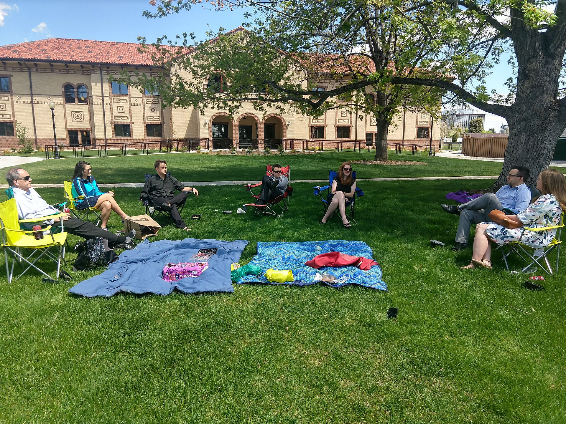 Seven people sitting on lawn chairs and blankets in a sunny park, with a brick building in the background. Relaxed and casual atmosphere.