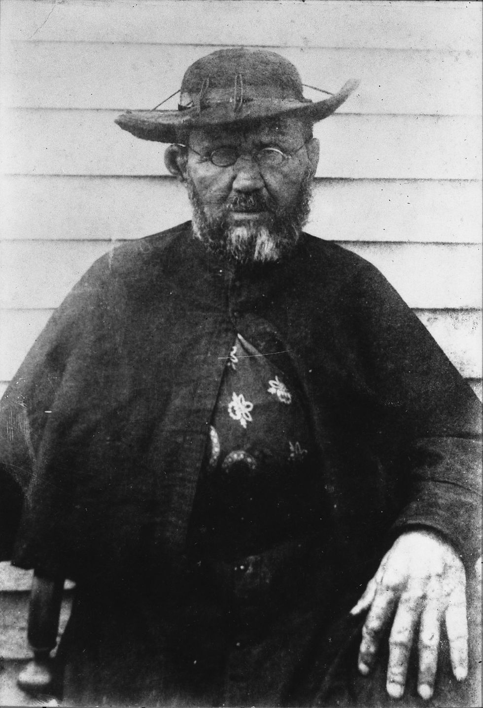 Black-and-white portrait of an older bearded man in a wide-brimmed hat and round glasses, standing solemnly before a clapboard wall.