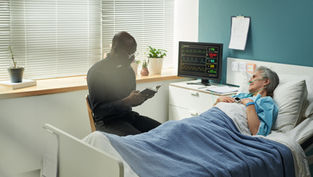 Elderly woman lies in hospital bed, connected to monitor. Clergyman reads beside her. Calm room with plants and blinds.